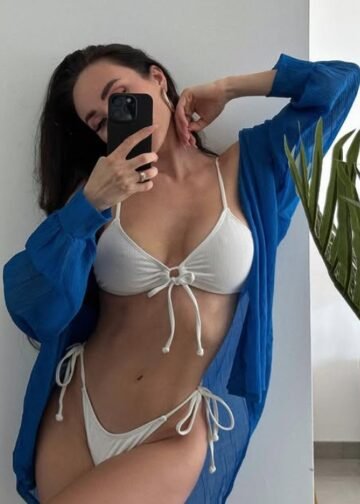 Escort Nyna wears a white bikini and a blue open shirt while taking a mirror selfie indoors in Hotel Amsterdam.